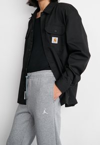 Černá košilová košile s dvěma kapsami na hrudi, nošená přes černé tílko, v kombinaci se šedými tepláky s bílým logem.