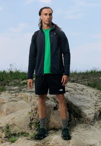 Completo da uomo per attività all'aperto che include una giacca in pile blu scuro con zip, una t-shirt verde, pantaloni corti neri, calze grigie e scarpe nere con accenti verdi.