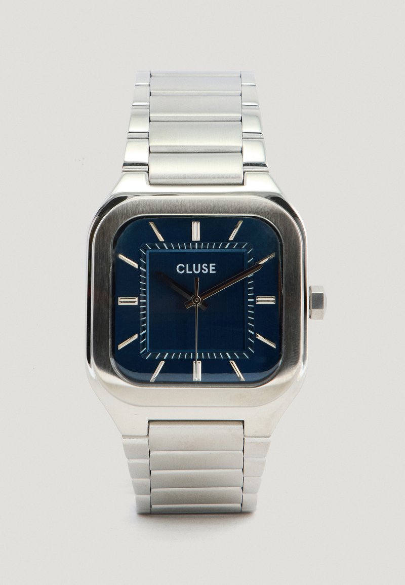 Montre carrée en acier inoxydable avec un cadran bleu, des index en argent et un bracelet en métal assorti. Marque Cluse affichée sur le cadran.