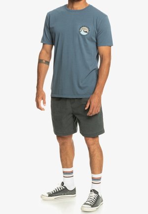 Camiseta de algodón azul con cuello redondo y un logo circular en el pecho. Combinada con pantalones cortos gris oscuro y zapatillas grises con cordones blancos.