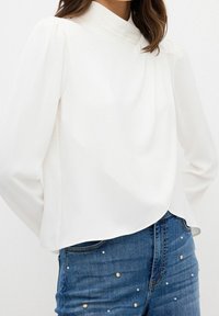 Femme portant un chemisier blanc à manches longues avec un col montant et un jean bleu orné de petites perles.