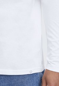 White long-sleeve shirt with a round neck, made of soft fabric, featuring a small circular logo tag at the hem. Paired with blue denim.