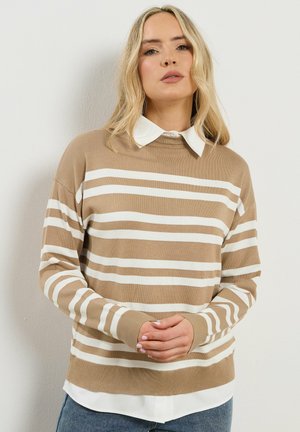 Blonde Frau, die einen beige-weiß horizontal gestreiften Pullover über einem weißen Hemd mit Kragen trägt, mit locker verschränkten Händen und vor einer schlichten Wand stehend.