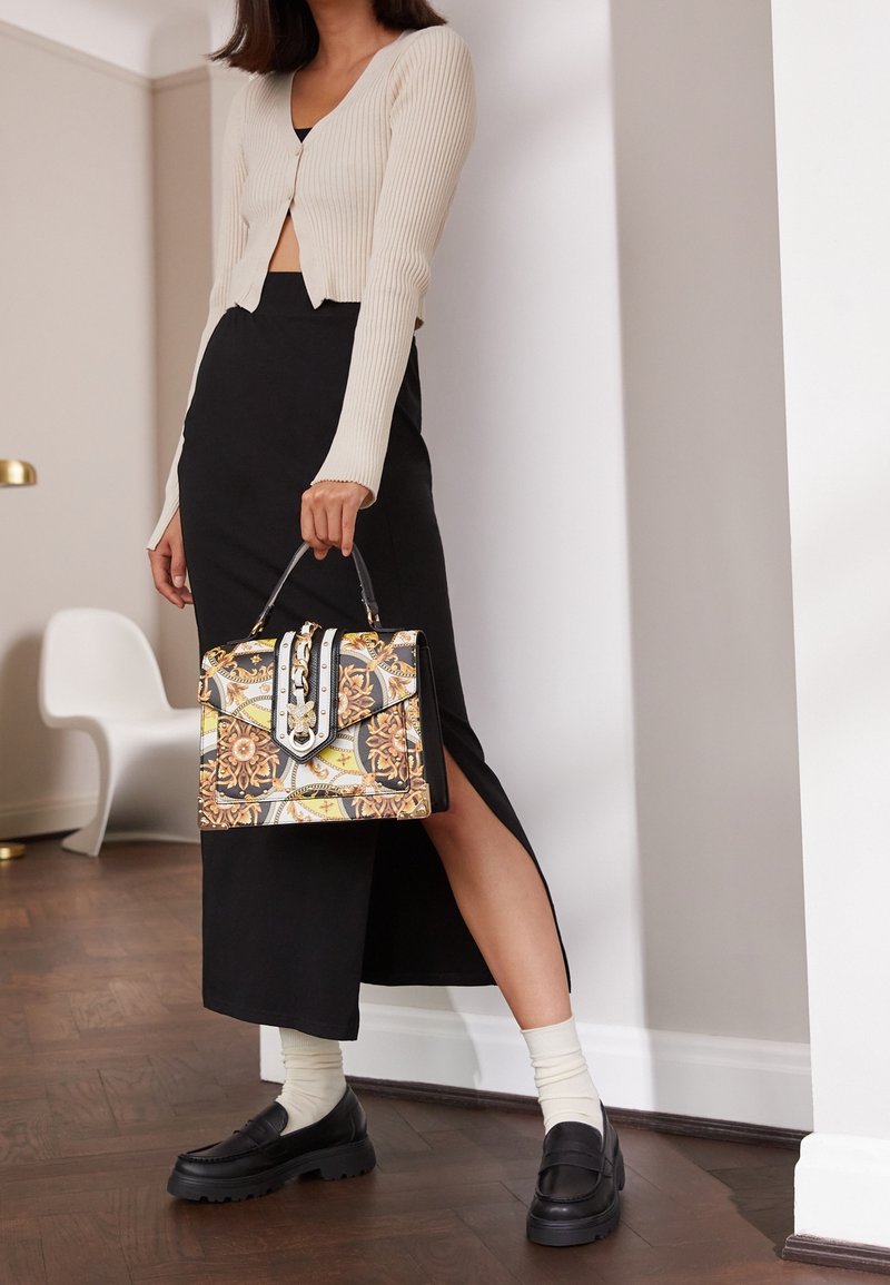 Femme en cardigan crème et jupe noire tenant un sac à main à motifs, portant des chaussettes blanches et des mocassins noirs à l'intérieur.