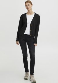 Čierny pletený cardigan s výstrihom do V, zapínaním na gombíky a rebrovanou textúrou, nosený cez biele tričko a skombinovaný s čiernymi nohavicami a teniskami.