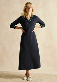 Navyblaues Langarmkleid mit V-Ausschnitt, grünen und weißen Akzenten und einer fließenden Silhouette. Kombiniert mit weißen Slippern mit einer Kettenverzierung.