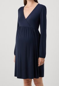 Námořnické modré těhotenské šaty s dlouhými rukávy, V- výstřihem a nařaseným materiálem pod prsy, mají volný střih a hladkou texturu.