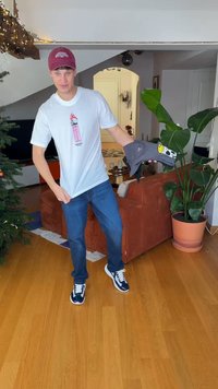 T-shirt blanc avec un motif rose, veste grise avec un visage souriant, jean bleu et baskets bleu marine. Cadre intérieur avec une plante et un canapé.