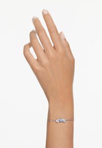 Pulsera de cadena de plata que presenta tres piedras preciosas: dos transparentes y una azul claro, con un diseño sencillo en una muñeca.