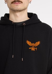 Homme moustachu portant un sweat à capuche noir avec un logo d'aigle orange tenant une clé sur la poitrine, cordons noirs et œillets en cuivre.