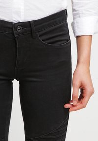 Jeans de mezclilla elástica negra con corte slim, que cuentan con un bolsillo frontal, cierre de botón y detalles de panel texturizado en la parte baja de las piernas.