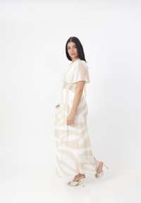 Robe longue et fluide en crème avec des motifs beige ondulés, manches courtes et taille smockée. Associée à des sandales à talons texturés et à bout ouvert.