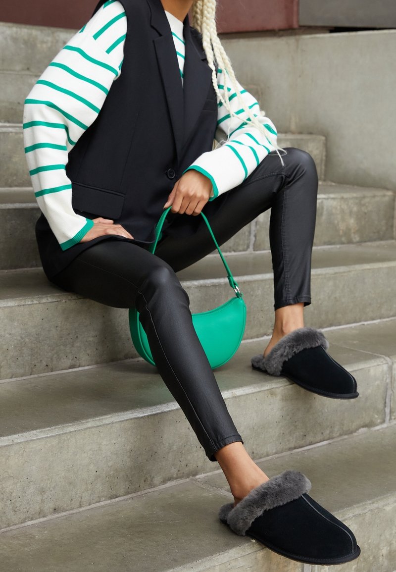 Personne assise sur des marches en béton portant un gilet noir, une chemise à manches longues rayée, des leggings noirs, des chaussons en fourrure grise, tenant un sac à main vert.