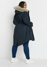 Marinblå parka-jacka med en huva klädd i fuskpäls, åtsittande midja och slitsar bak. Kombinerad med blå jeans och vita sneakers.