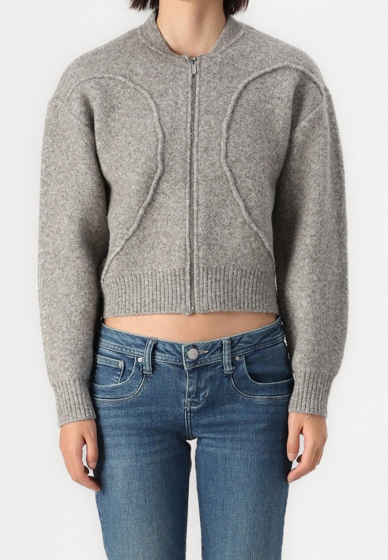 Femme portant un pull court gris zippé avec des détails de coutures en relief, associé à un jean bleu, debout devant un fond uni.