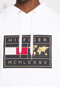 Hvid hættetrøje med et stort logo-patch i marineblå og guld, der viser et verdenskort, en rød rektangel og teksten "HILFIGER". Snore med guldtipper.