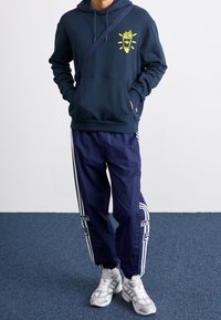 Námořnický hoodie s žlutou grafikou, přední klokaní kapsa; odpovídající námořnické sportovní kalhoty s bílými bočními pruhy a logem, doplněné šedými teniskami.