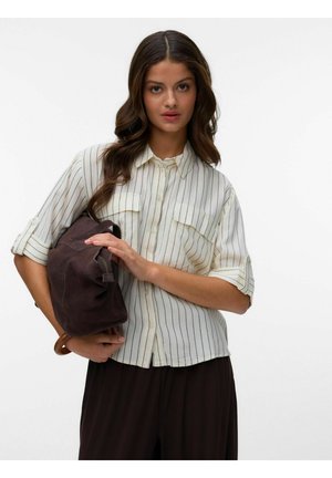 Žena s dlhými hnedými vlasmi, oblečená v bielej pásikavej košeli na gombíky a čiernych nohaviciach, drží tmavohnedú koženú tašku.