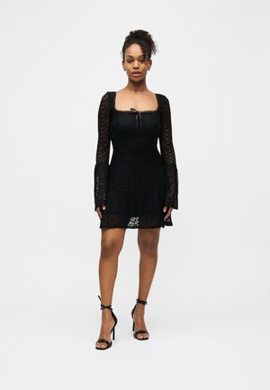 Frau trägt ein schwarzes Spitzen-Minikleid mit langen Glockenärmeln und quadratischem Ausschnitt, kombiniert mit schwarzen, hochhackigen Sandalen mit Knöchelriemen.