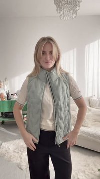 Junge Frau mit blonden Haaren, die eine hellgrüne gesteppte Weste über einem beigen Hemd trägt, steht in einem hellen Wohnzimmer mit grünem Schrank und Kronleuchter.