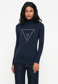 Mujer con pelo largo y oscuro que lleva un suéter de cuello alto de Guess en azul marino con un logo brillante y jeans azul oscuro, de pie contra un fondo blanco.