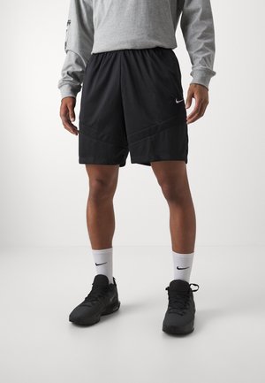 Persona con pantalones cortos deportivos negros de Nike, calcetines blancos de Nike, zapatos deportivos negros y una camiseta de manga larga gris, de pie sobre un suelo blanco.