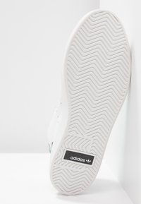 Bílá podrážka tenisky Adidas s rybí kostí vzorem běhounu a černým logem Adidas blízko paty, na bílém pozadí.