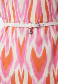 Weißer Ledergürtel mit einer runden silbernen Schnalle und Charm, getragen über einem Stoff mit einem Muster in Rosa, Orange und Creme, das Ikat ähnelt.