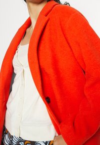 Donna con un cappotto di lana arancione brillante sopra un cardigan crema con bottoni e pantaloni a fantasia, mano in tasca, su sfondo bianco.