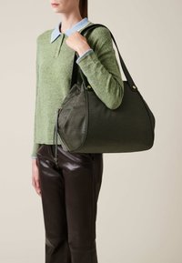 Une personne tient un sac à main texturé vert foncé avec des poignées noires, portant un pull vert avec un col bleu et un pantalon en cuir noir.