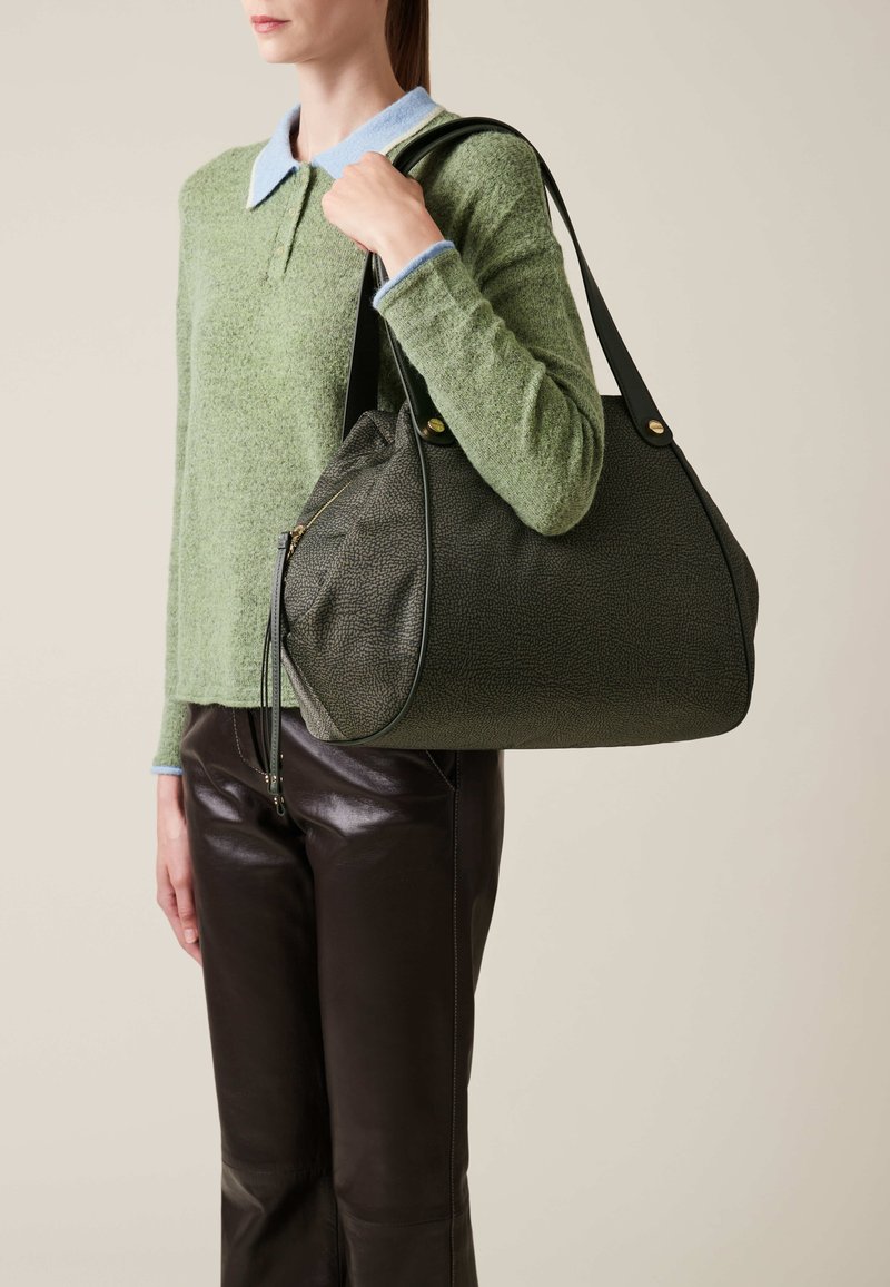 Une personne tient un sac à main texturé vert foncé avec des poignées noires, portant un pull vert avec un col bleu et un pantalon en cuir noir.