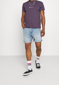 Fialové bavlněné tričko s bílým logem, džíny šortky s ohrnutými rukávy, černé tenisky a bílé ponožky s červenými pruhy.