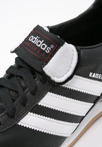 Zwarte leren sneaker met drie witte strepen, textuur oppervlak en een zwart-wit merk-velcroband bij de enkel.
