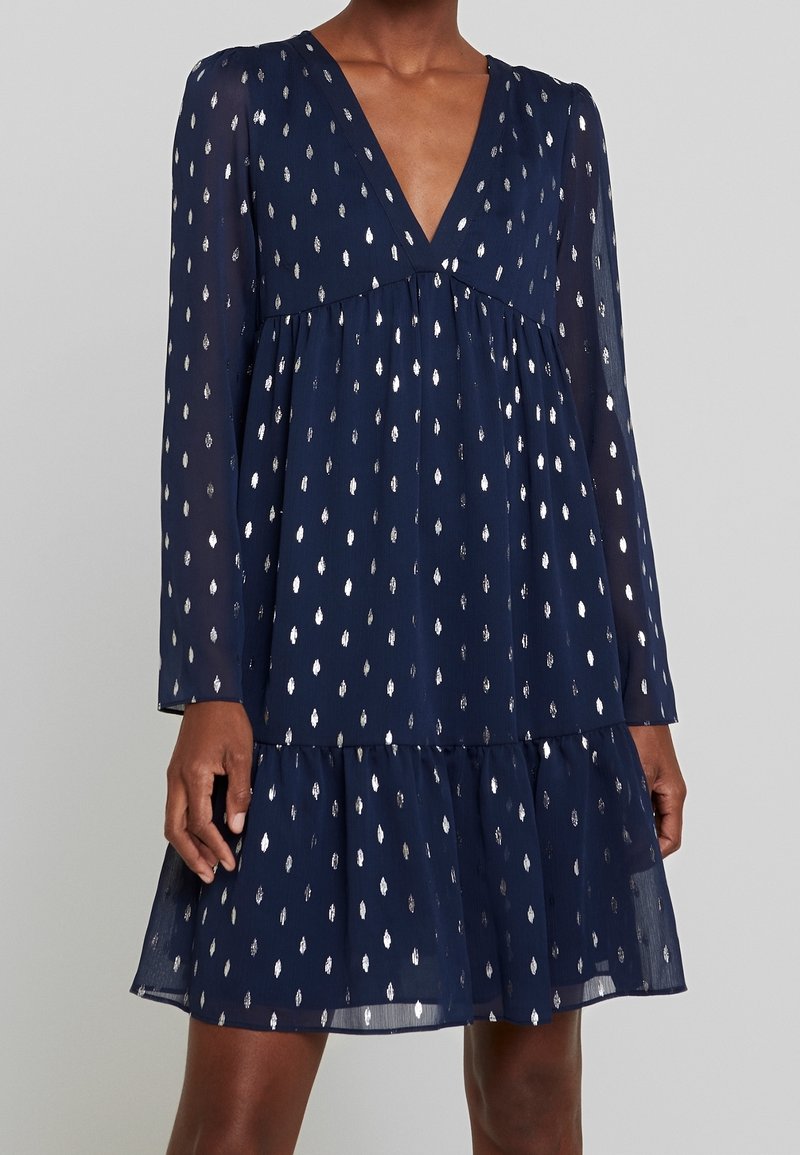 Marineblauwe jurk van doorschijnende stof, met een V-hals, lange mouwen en een zilver gestippeld patroon. Uitlopende, gelaagde zoom die beweging toevoegt.