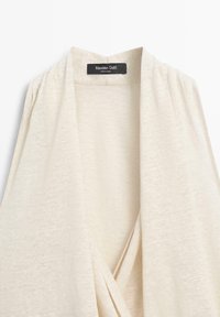 Helles, beiges Cardigan mit offenem Vorderteil und weicher, gestrickter Textur sowie einem schwarzen Massimo Dutti Etikett, das innen am Halsausschnitt eingenäht ist.