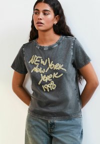 Tumši pelēks T-krekls ar apaļu kaklu un īsām piedurknēm, kurā ir spīdīgi zeltīti uzraksti: "NEW York 1998" dekoratīvā stilā.