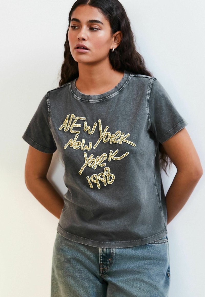 Mørkegrå T-shirt med rund hals og korte ærmer, prydet med skinnende guldbogstaver: "NEW York 1998" i en dekorativ stil.