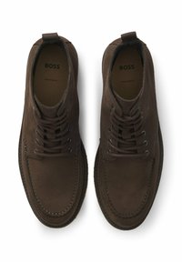 Brązowe zamszowe buty o zaokrąglonym nosku, z zapięciem na sznurowadła, szczegółami szwów i pętelką na pięcie. Wewnątrz znajduje się logo marki.