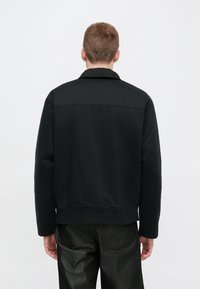 Fekete pamut dzseki galléros nyakkal, bordázott szegéllyel és minimalista kialakítással. Simább textúra, a hátulnézet tiszta vonalakat és varrást mutat.