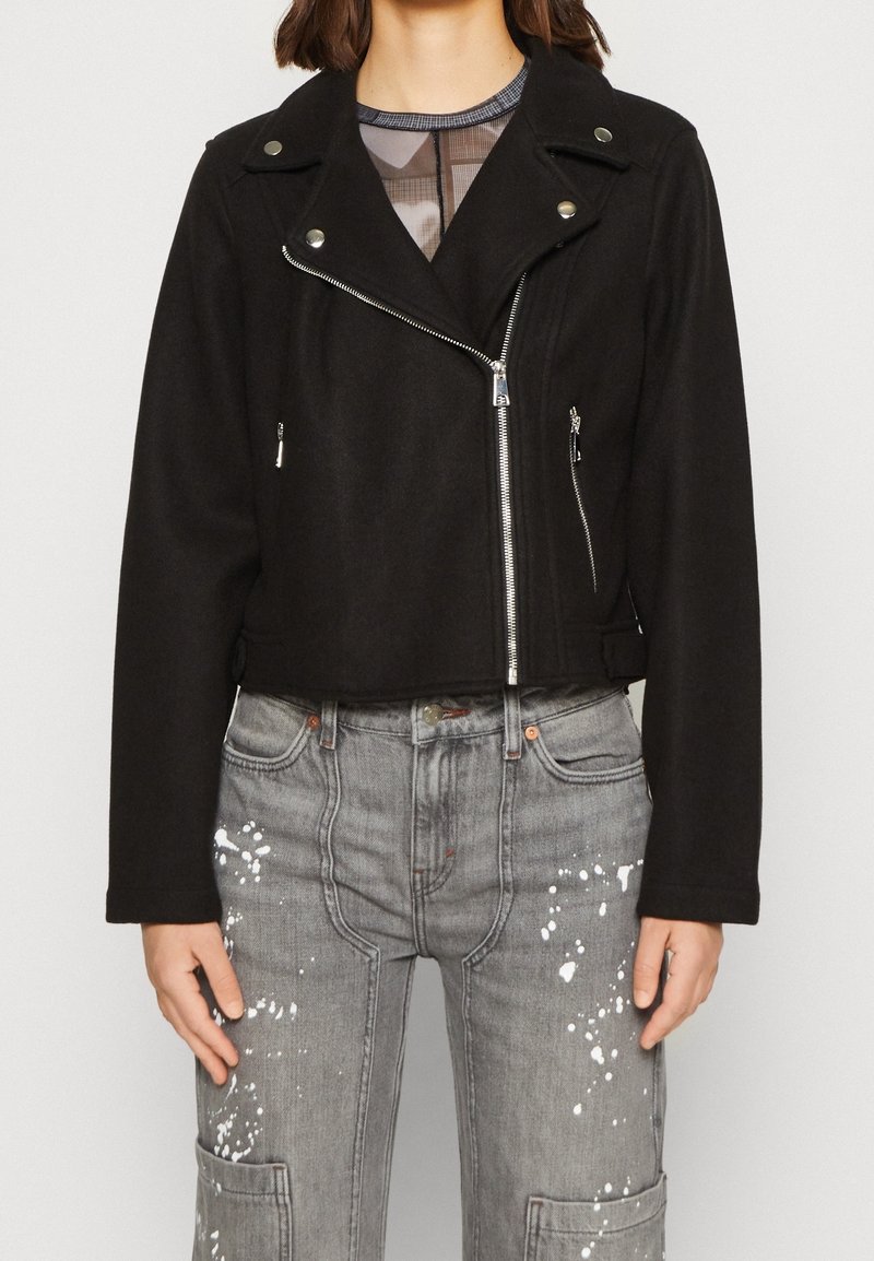 Femme portant une veste courte de motard noire avec des fermetures éclair argentées sur un top transparent, associée à un jean cargo gris éclaboussé de peinture.