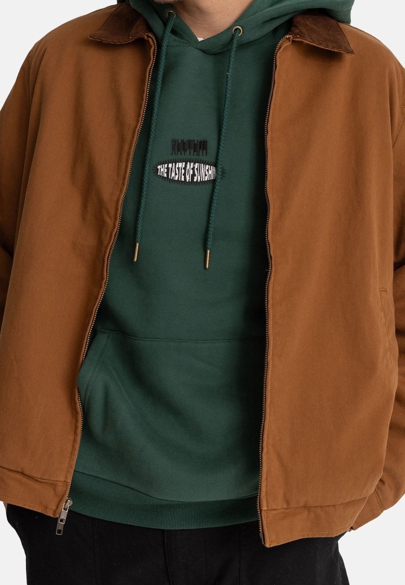 Ruskea takki, jossa samettinen kaulus, ja vihreä huppari alla. Hupparissa on graafinen tekstikuvio ja kultaiset nauhanpäätteet.