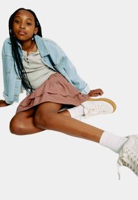 Jeansjacka, grå henley-tröja, lager av rosa kjol med prickigt mönster, och vita sneakers med texturerad sula; sittande pose mot en vit bakgrund.