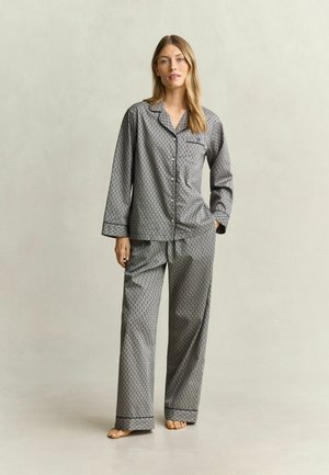 Frau steht barfuß und trägt einen passenden grauen Pyjama mit Knöpfen, langen Ärmeln und kleinem schwarzem geometrischem Muster.