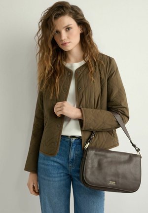 Młoda kobieta o długich falowanych włosach nosi oliwkową pikowaną kurtkę i niebieskie dżinsy, trzyma ciemnobrązową skórzaną torbę na ramię ze złotym logo.