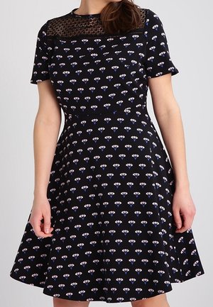 Vrouw draagt een zwarte jurk met korte mouwen met kleine paarse en witte bloemenprints en een zwart kanten inzetstuk op de bovenborst.