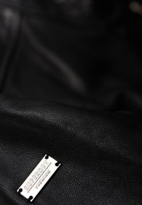 Tessuto in pelle nera con una superficie texturizzata e un'etichetta in metallo argentato che riporta la scritta "SUPERDRY PREMIUM" attaccata.