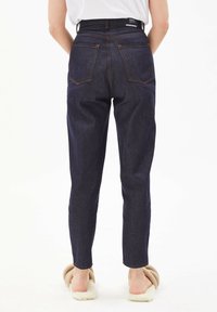 Pantalon en denim bleu foncé avec une silhouette ajustée, taille haute et deux poches arrière, présentant des coutures contrastantes orange et des jambes fuselées.