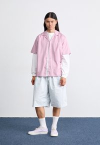 Ein hellrosa T-Shirt mit kurzen Ärmeln über einem weißen Langarmshirt, kombiniert mit hellblauen Shorts und pastelllila Turnschuhen. Einfaches Design.