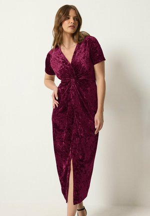 Donna in un abito di velluto bordeaux scuro a maniche corte, con drappeggio frontale e spacco, in piedi davanti a uno sfondo chiaro e semplice.