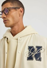 Jasno beżowa kurtka z granatowym haftowanym "N" na piersi, noszona na jasno kremowym hoodie. Posiada zatrzaski i gładką teksturę.